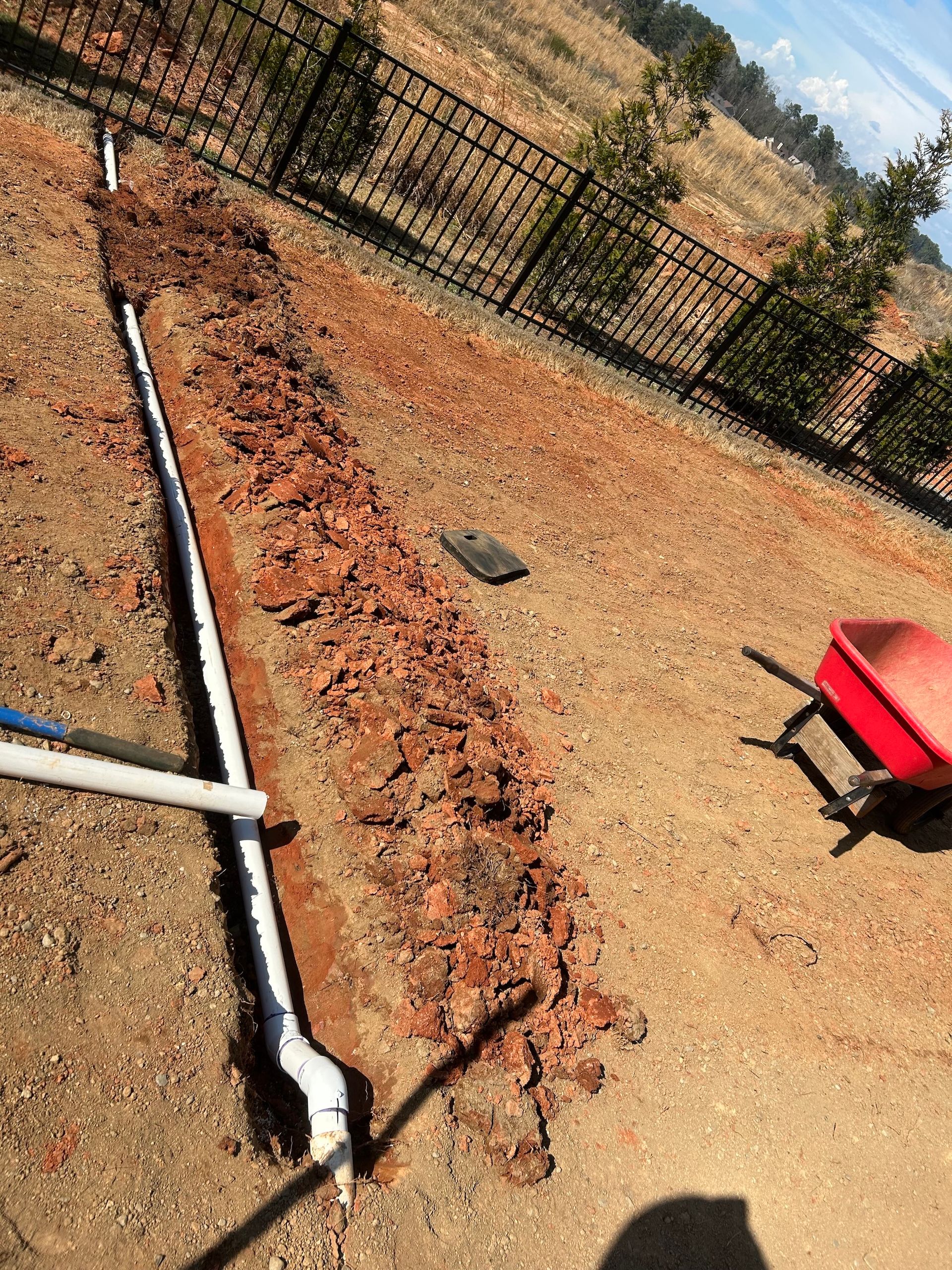 Dry backyard with a long black hose, brick border, fence, and a red wheelbarrow on gravel.
