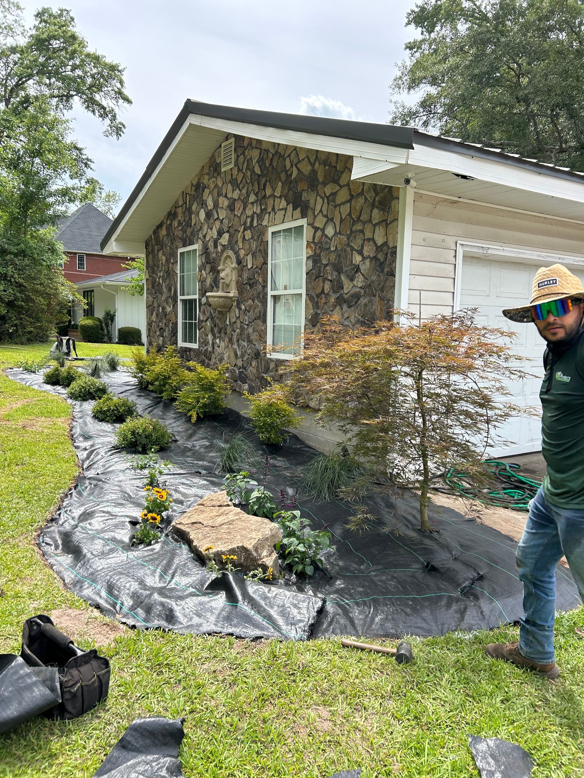 Stone-sided house with fresh black landscaping fabric, shrubs, and a worker standing in the yard.