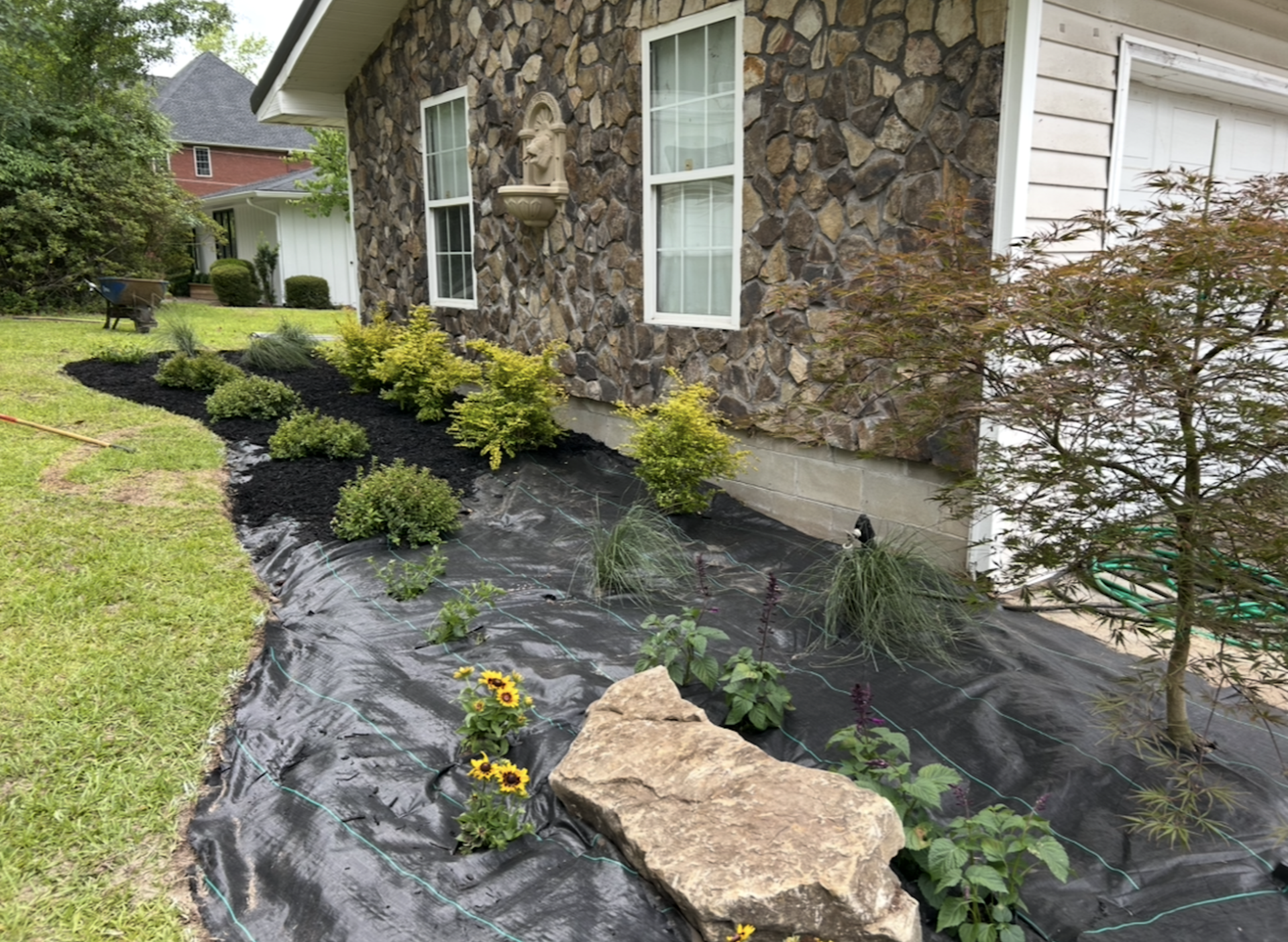 Landscaped garden bed with black mulch, shrubs, flowers, and a stone beside a stone house