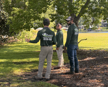 Three people in green jackets stand talking in a sunlit park with trees and grass.