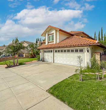 Two-story suburban house with red tile roof, white garage, driveway, and green lawn under a partly cloudy sky