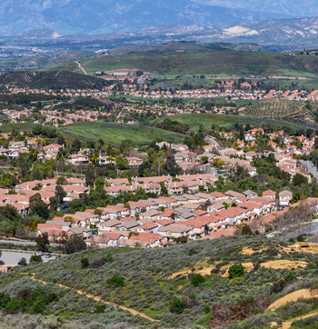 A hillside neighborhood with terracotta-roofed homes and green fields, overlooking distant mountains.