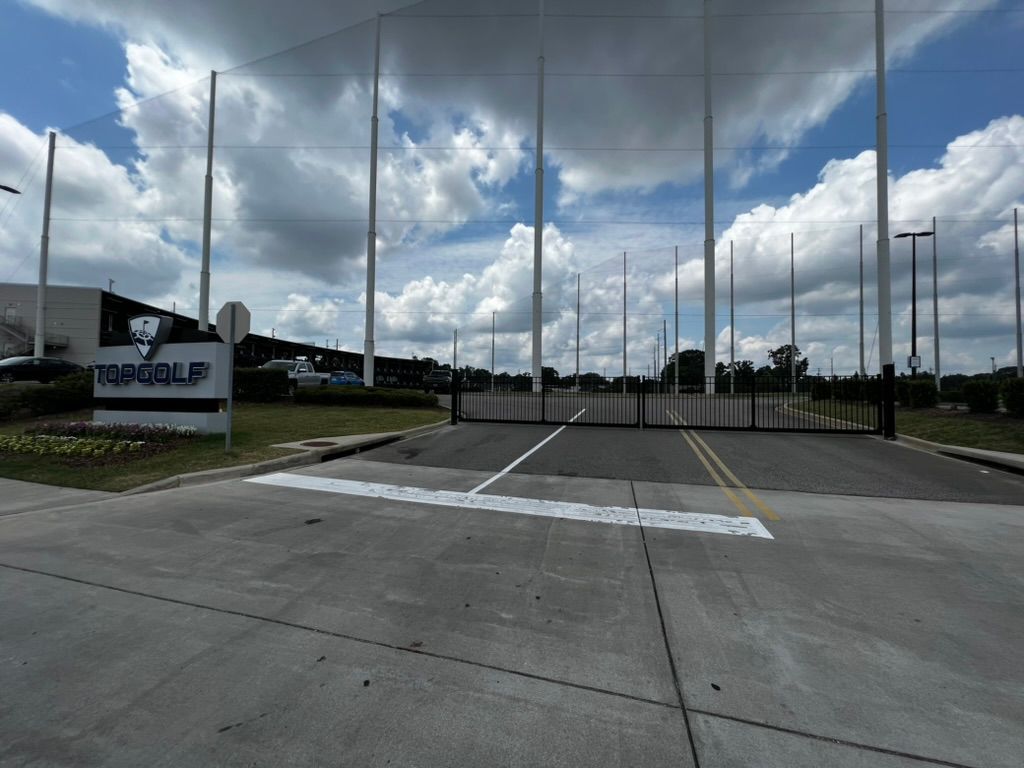 Entrance to a Topgolf facility with tall netting, sign, and cloudy sky.
