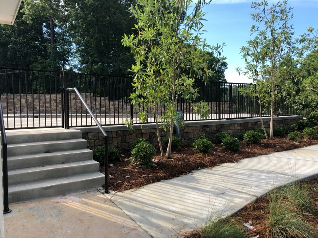 Concrete steps with railing lead to a walkway alongside a black metal fence and landscaped area with trees.