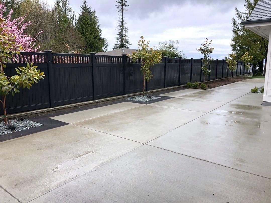 Black fence bordering a concrete driveway with small trees and landscaping; cloudy sky.