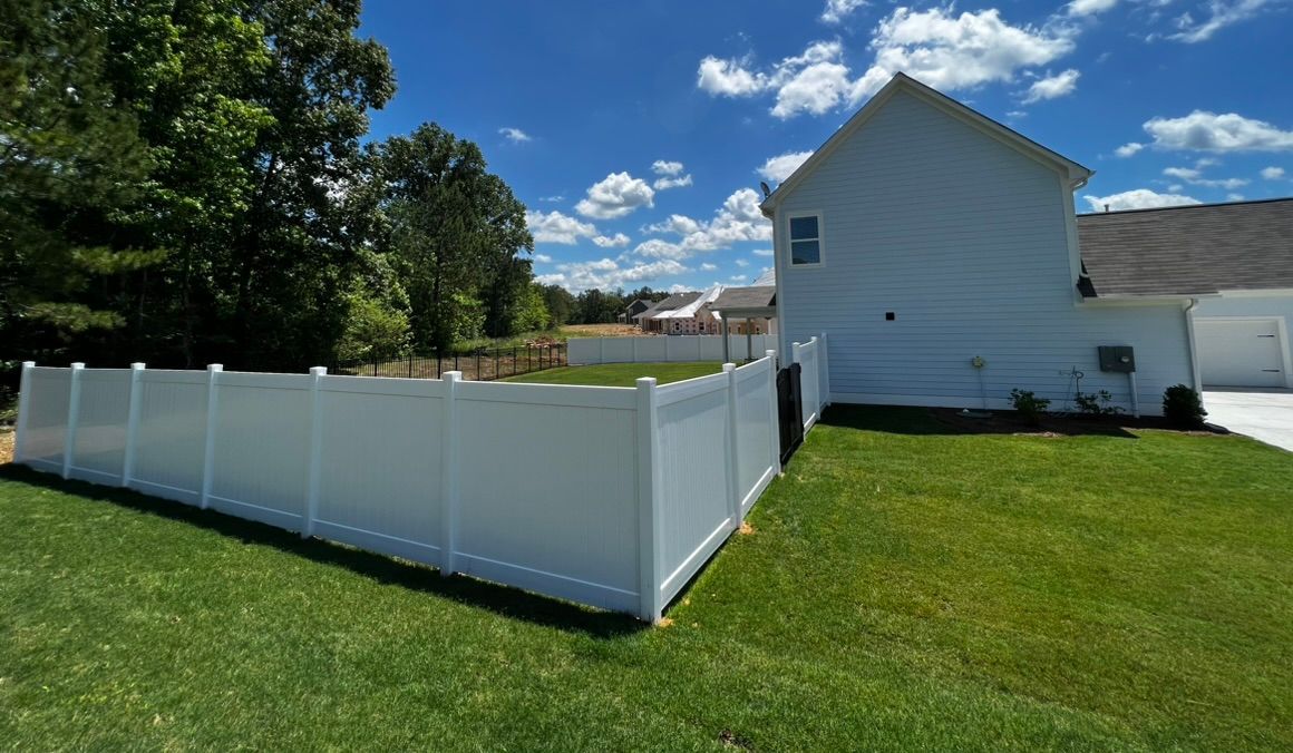 White vinyl fence surrounds a green lawn and a white house under a blue sky with clouds.