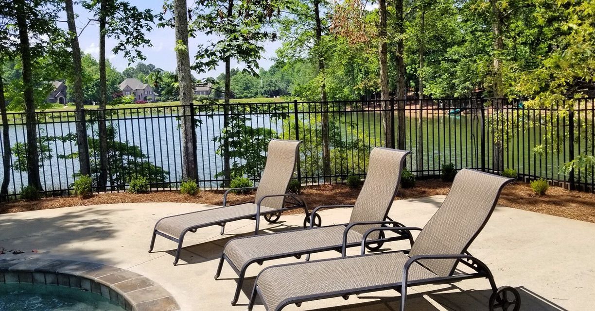 Three lounge chairs on a patio overlooking a lake behind a black fence, surrounded by trees.