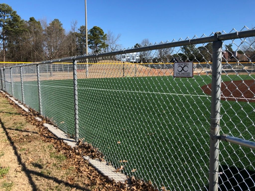 Chain-link fence surrounding a baseball field with a green turf. A sign on the fence reads 