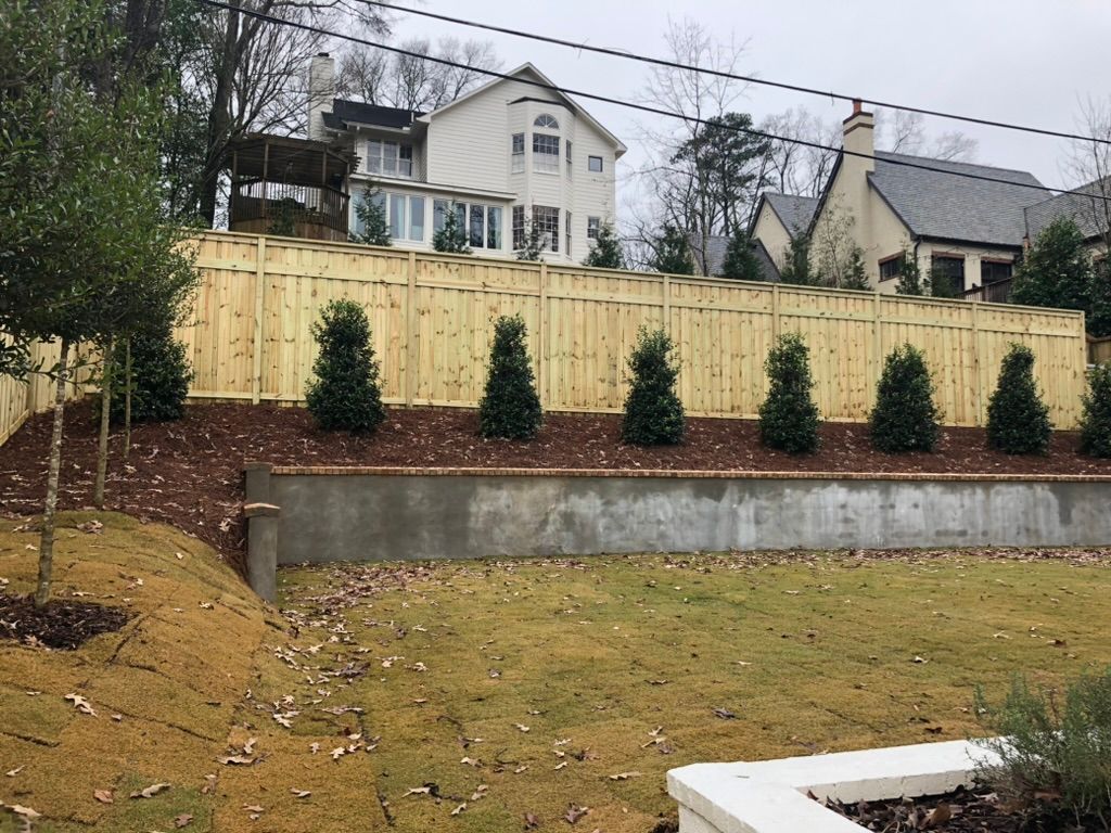 A wooden fence lines a yard with green grass and trees, with a house in the background.