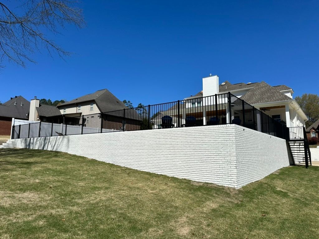 A brick retaining wall supports a raised deck with a black railing, house in background.