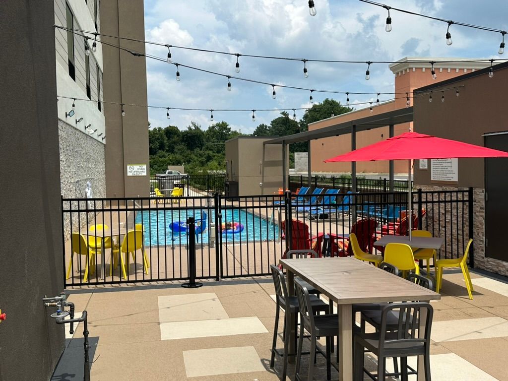 Outdoor patio with tables, chairs, and a pool. A red umbrella provides shade, and string lights hang overhead.