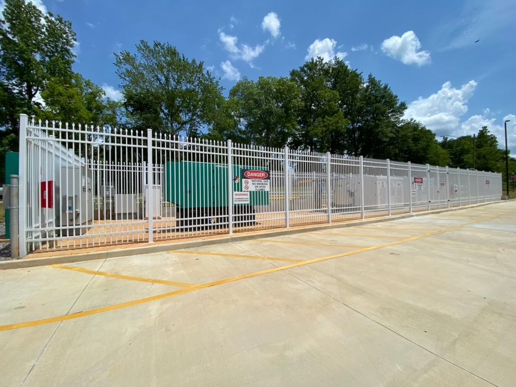 White fenced-in electrical equipment on concrete, with caution signs. Blue sky and trees in the background.
