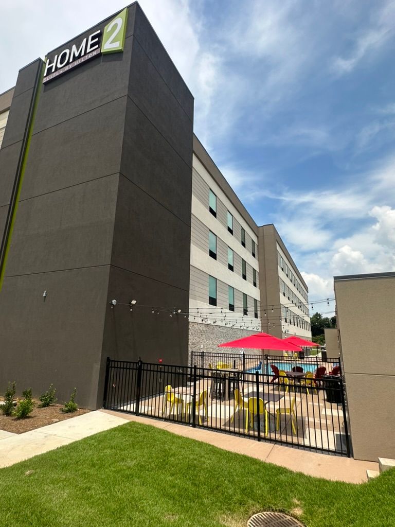 Home2 Suites hotel exterior with a pool area. Gray and white building with a black fence, red umbrella, and string lights.