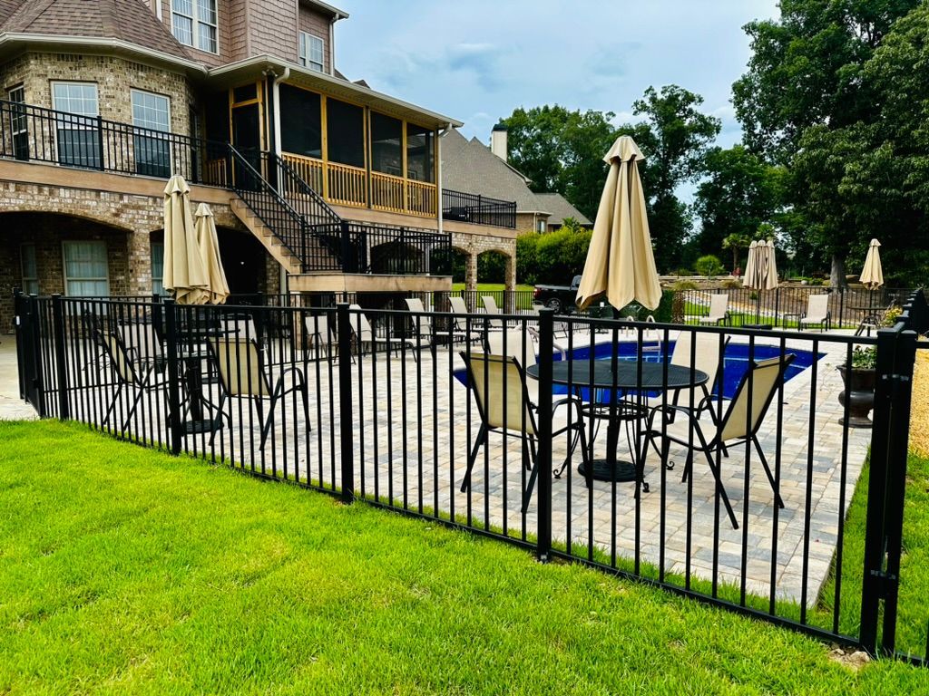 Black fenced patio with table, chairs, umbrellas, and a large house in the background. Green grass.