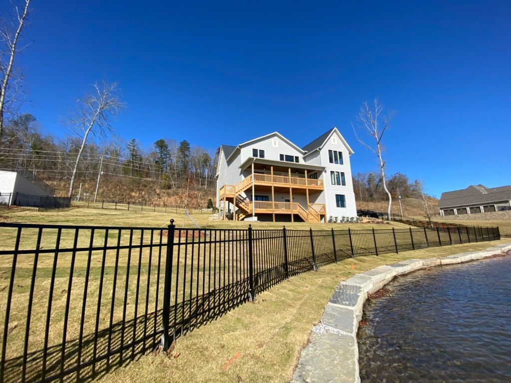 Two-story house with deck, black fence, and pond under a clear blue sky.