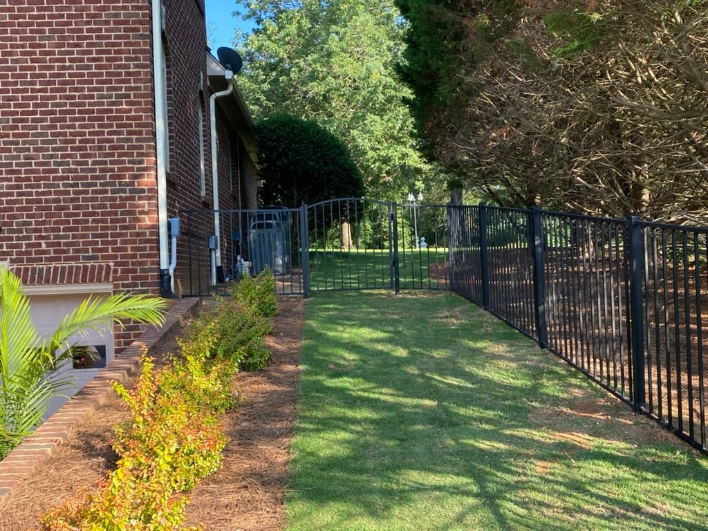 Black metal fence encloses a green lawn along the side of a brick building and a line of bushes.