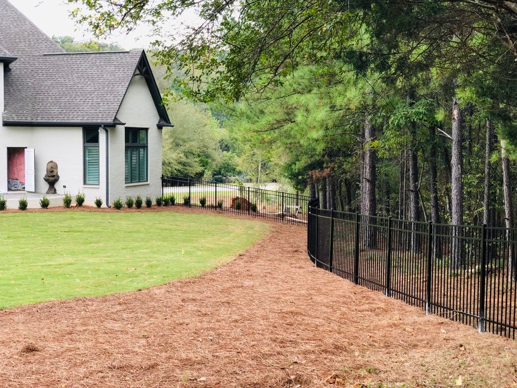A modern house with a green lawn and a path covered in mulch next to a black fence and trees.