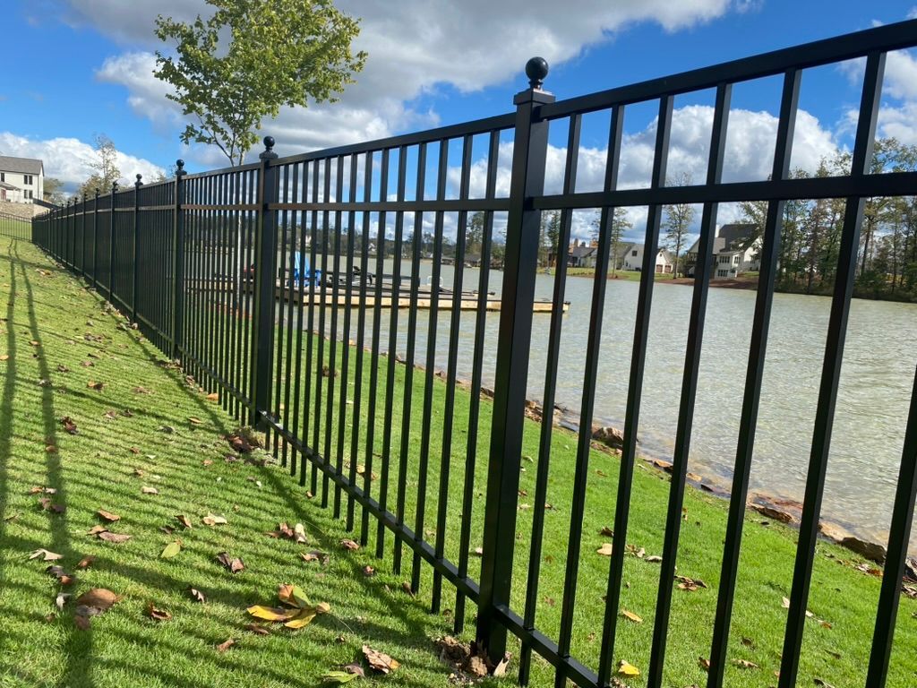 Black metal fence bordering a green grassy area beside a lake under a blue sky.