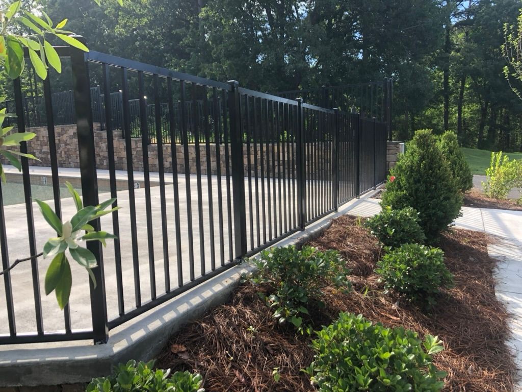Black metal fence bordering a pool area with a bed of mulch and green shrubs.