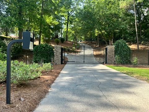 Gated driveway with black gates, brick pillars, and a gray asphalt road. Surrounded by trees and greenery.