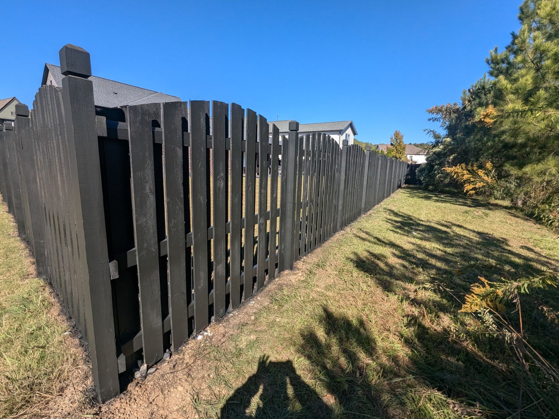 Black wooden fence in a grassy area under a blue sky, with houses in the background.