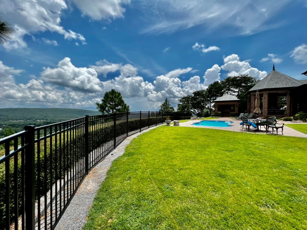 Lush green yard with black fence overlooks a scenic vista, with a pool and gazebo on the right, under a blue sky with clouds.