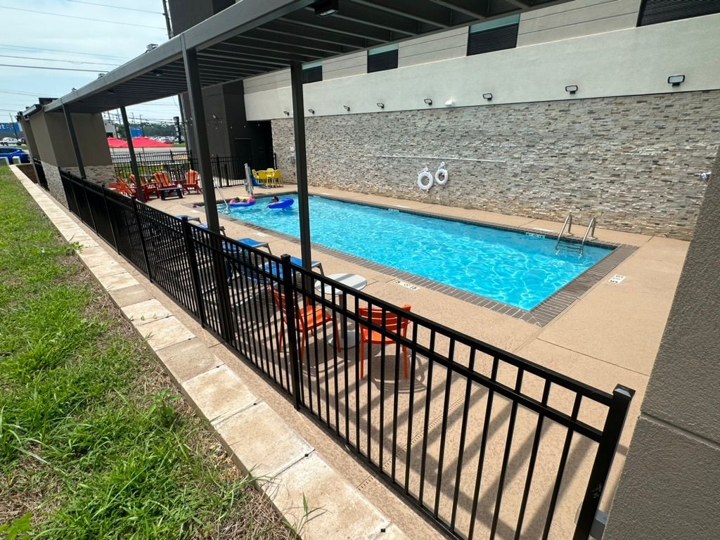 A black fenced-in rectangular pool with a stone wall and awning. Lawn and patio furniture are nearby.