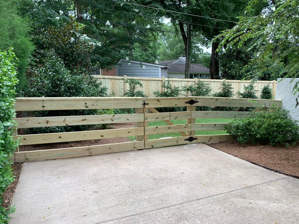 Wooden fence with two gates on a concrete patio, with green bushes and trees in the background.