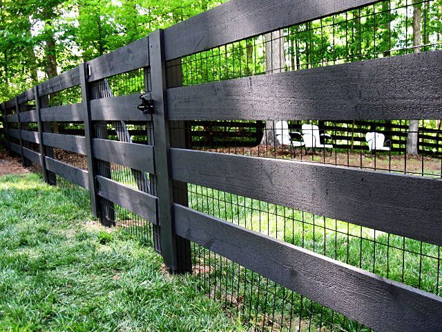 Black wooden fence with wire mesh, in a grassy area with trees in the background.