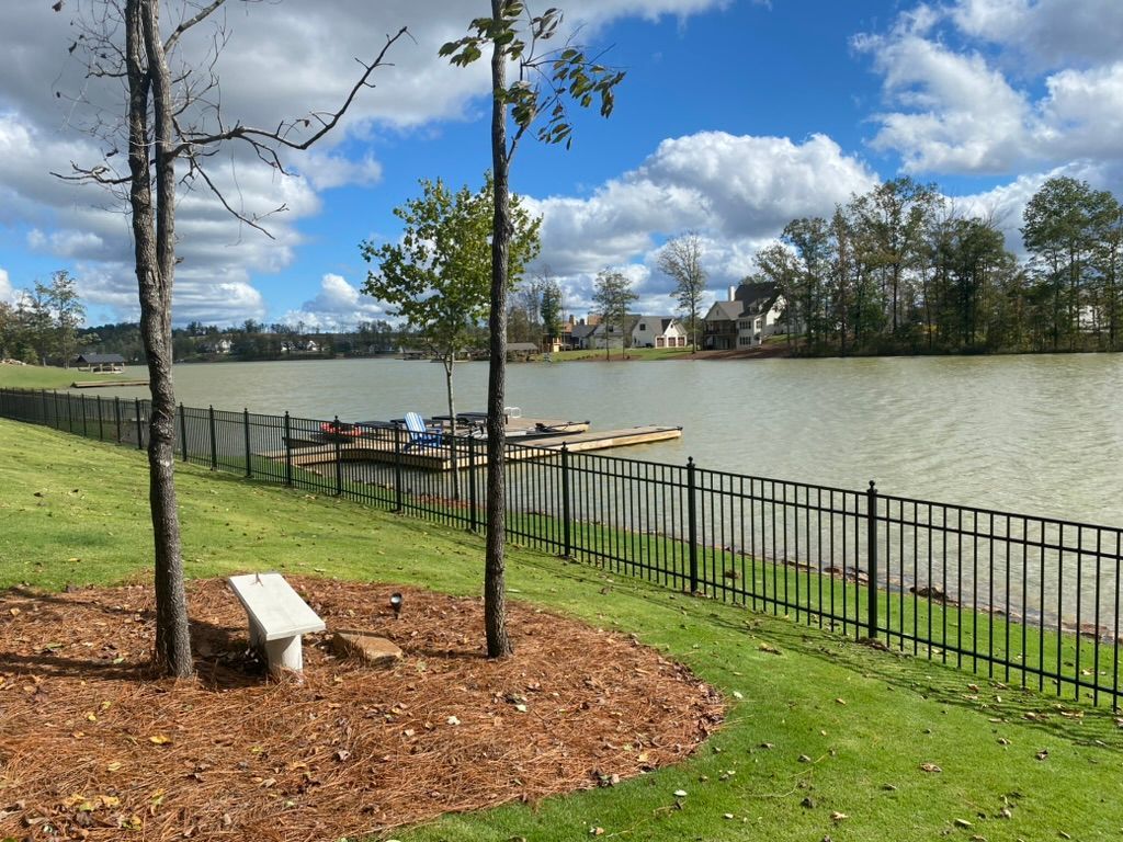 Lakeside view with a bench, trees, and black fence. Houses and cloudy sky in the background.