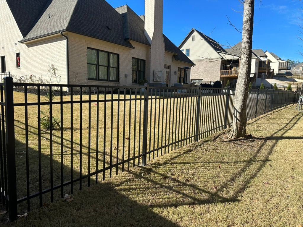 Black metal fence bordering a grassy yard in front of multiple houses under a blue sky.