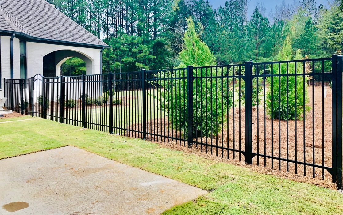 Black metal fence surrounds a grassy yard next to a beige patio and a house with a tiled roof.