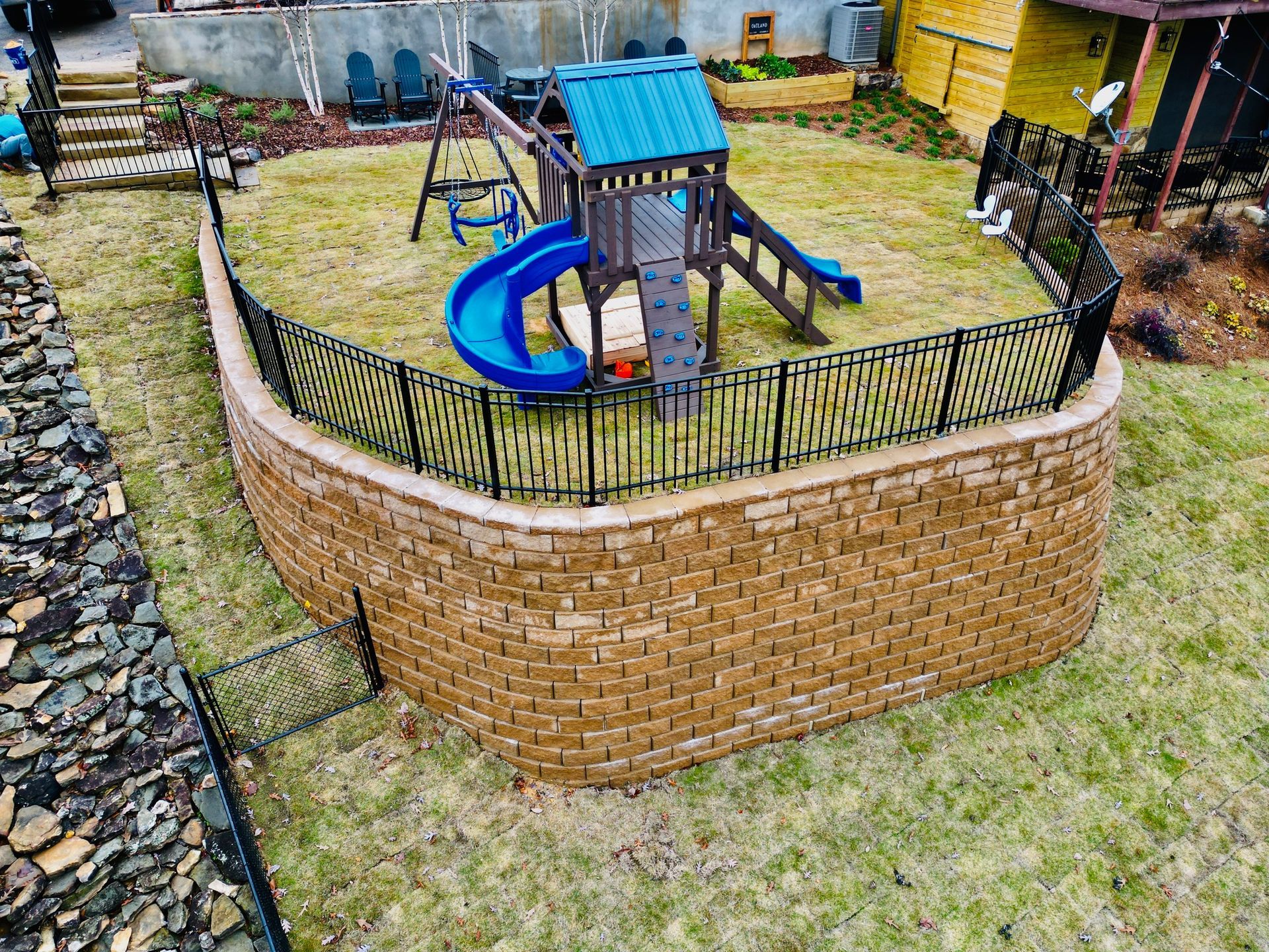 Playground enclosed by a retaining wall with black fence, on sloped yard with sparse grass.
