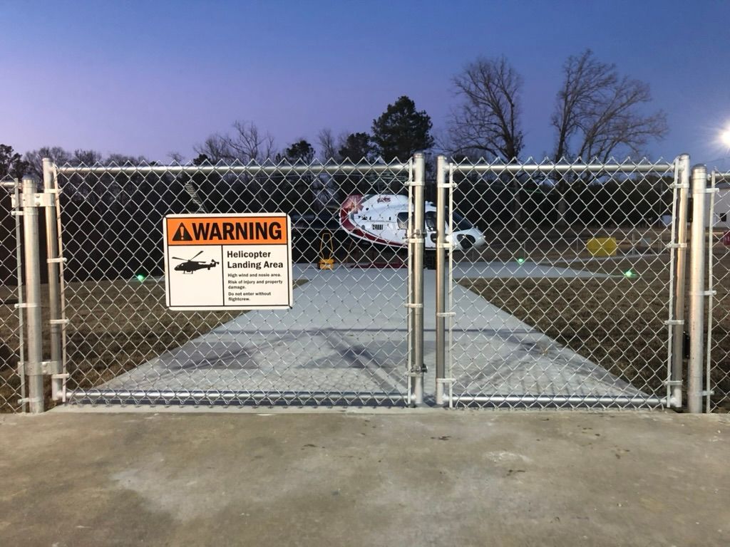 Chain-link fence with a warning sign, blocking access to a helicopter landing pad. Helicopter visible in the background.