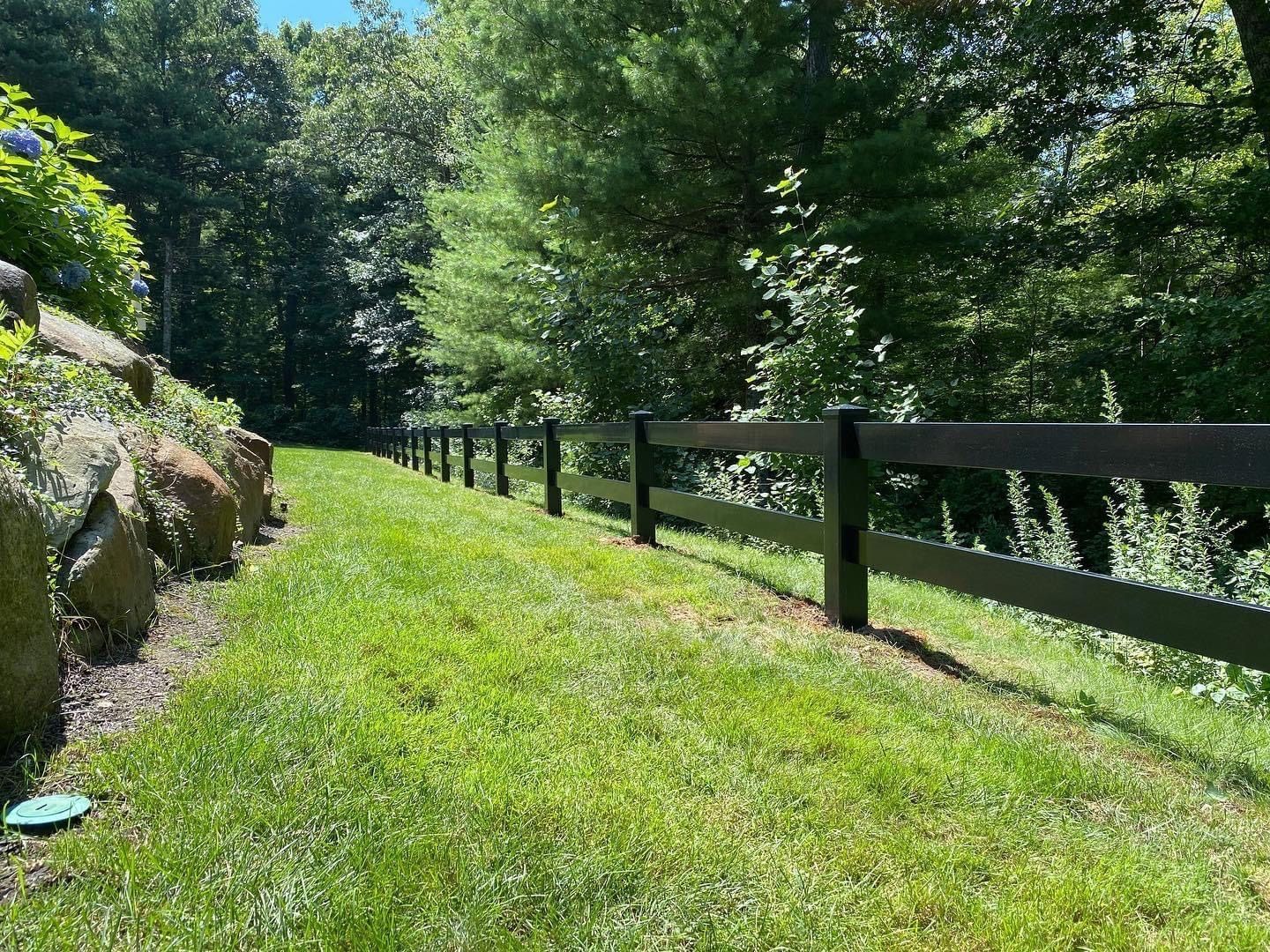 Black wooden fence on a grassy hillside, surrounded by trees.