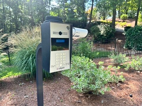 Metal security access panel with keypad and camera, mounted outdoors near shrubbery and trees.