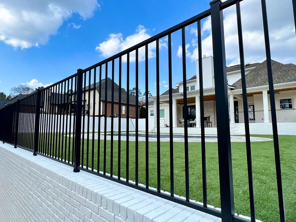 Black metal fence atop a white brick wall, in front of a green lawn and two-story house under a blue sky.