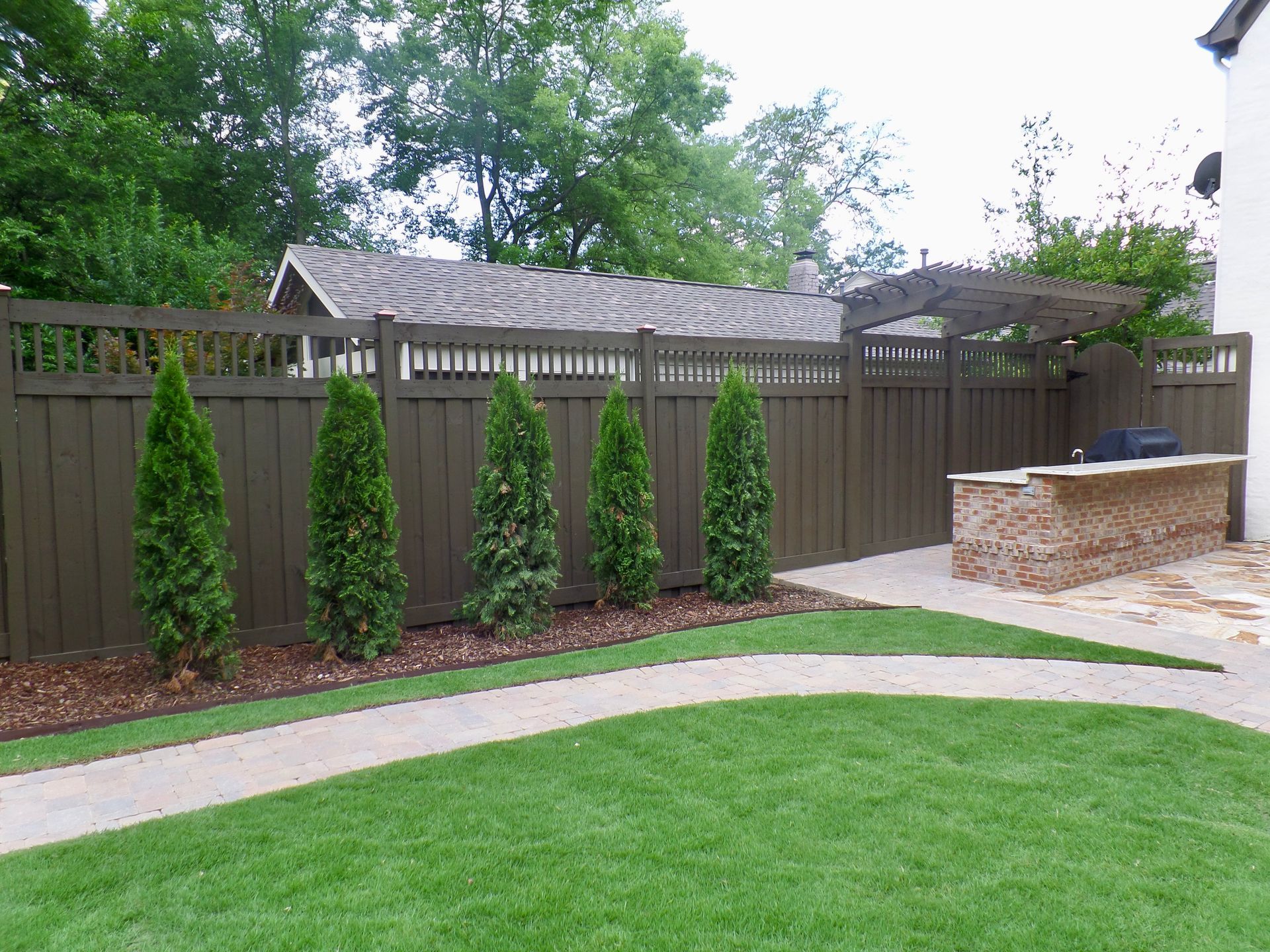 Green yard with a brown wooden fence, trees, and stone patio.