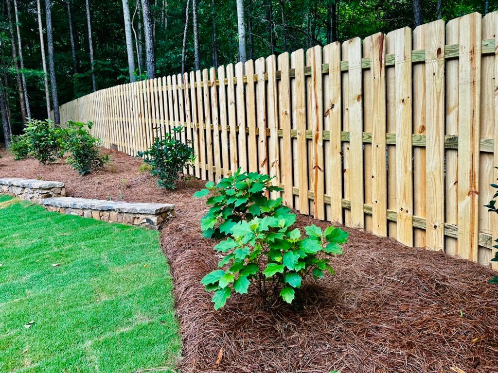 Wooden fence bordering a landscaped yard with green bushes, mulch, and a grassy lawn.
