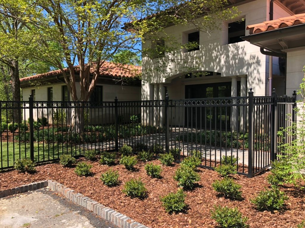 Black fence surrounds a building with a red-tiled roof and landscaping with mulch and shrubs.