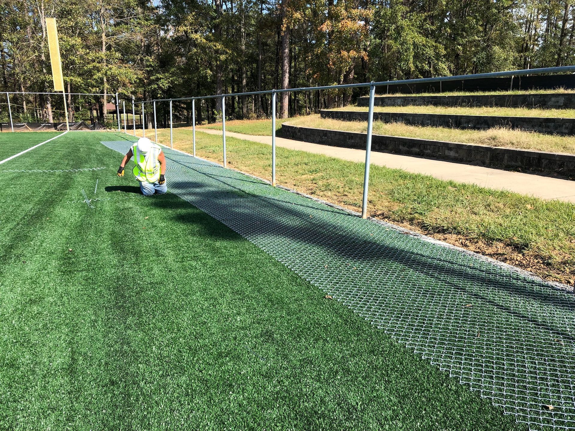 Person applying treatment to a green turf field near a metal fence and tiered seating.