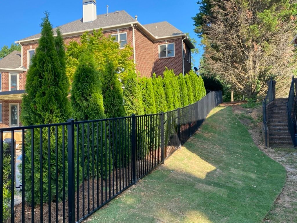 Black fence bordering a grassy hill next to a line of green trees and a brick house.