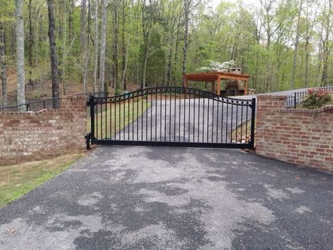 Black metal driveway gate on a brick wall, leading to a paved driveway and a wooden pergola.