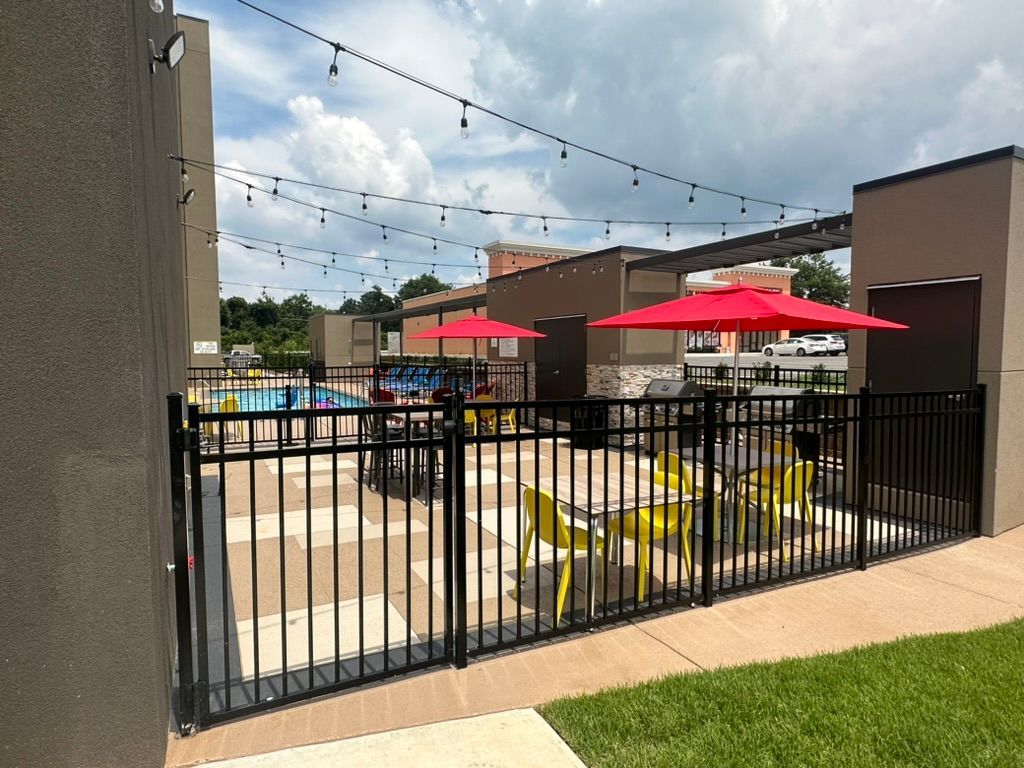 Outdoor patio with pool. Black fence surrounds yellow tables with red umbrellas, string lights overhead.