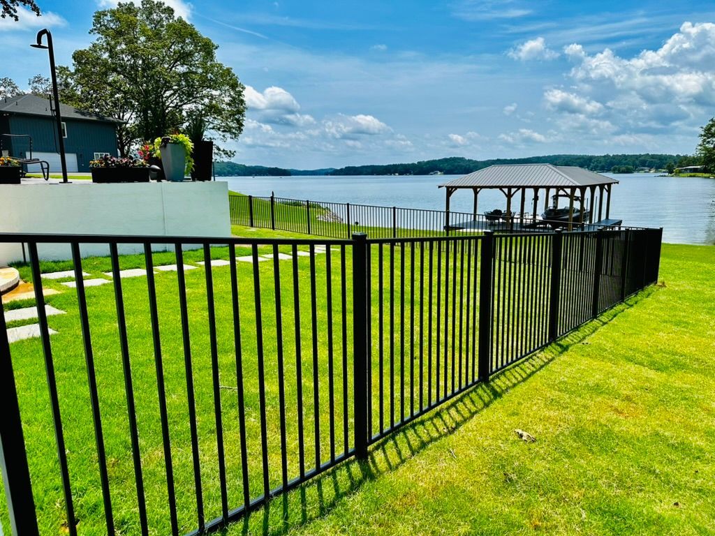 Black metal fence bordering a green lawn leading to a lake with a dock, under a blue sky.