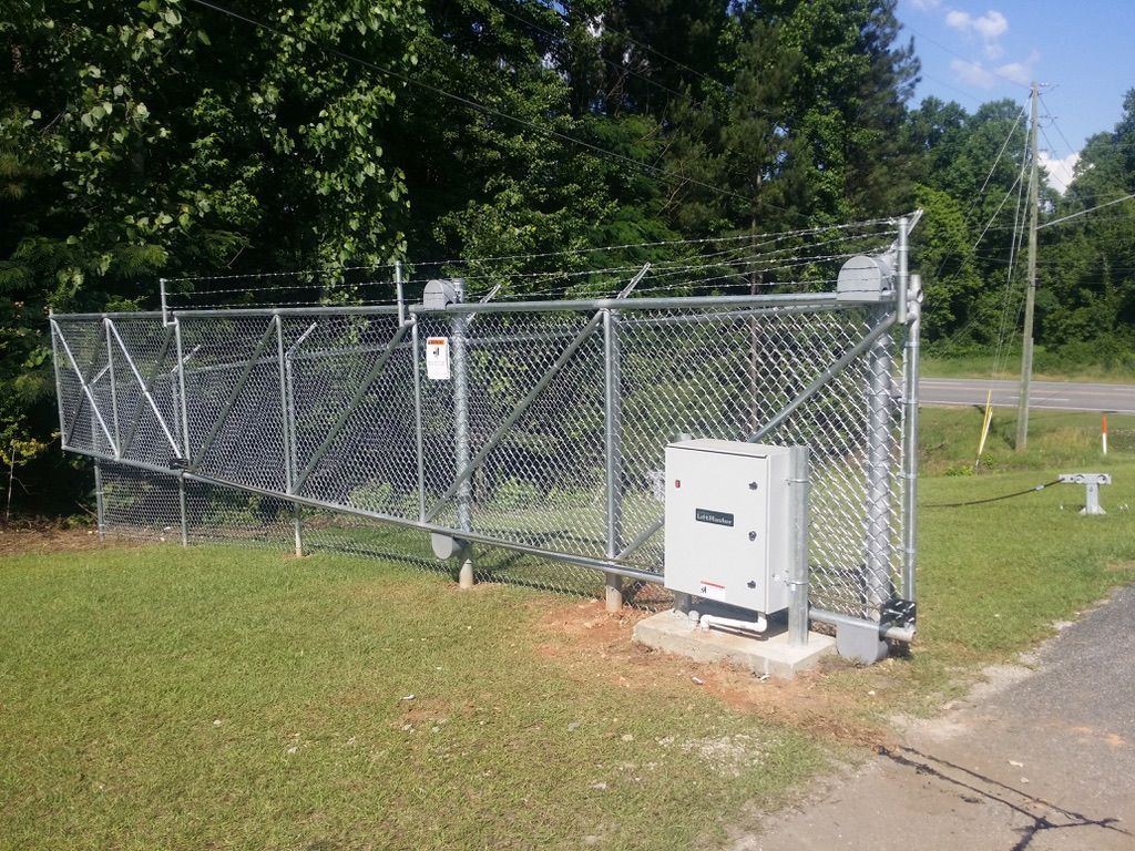 Chain-link fence with barbed wire surrounding a gray electrical box on a concrete pad, next to road and trees.