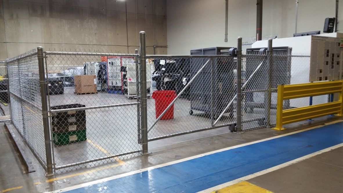Warehouse area with chain link fence enclosure. Gray containers, red object inside. Blue and yellow floor markings.