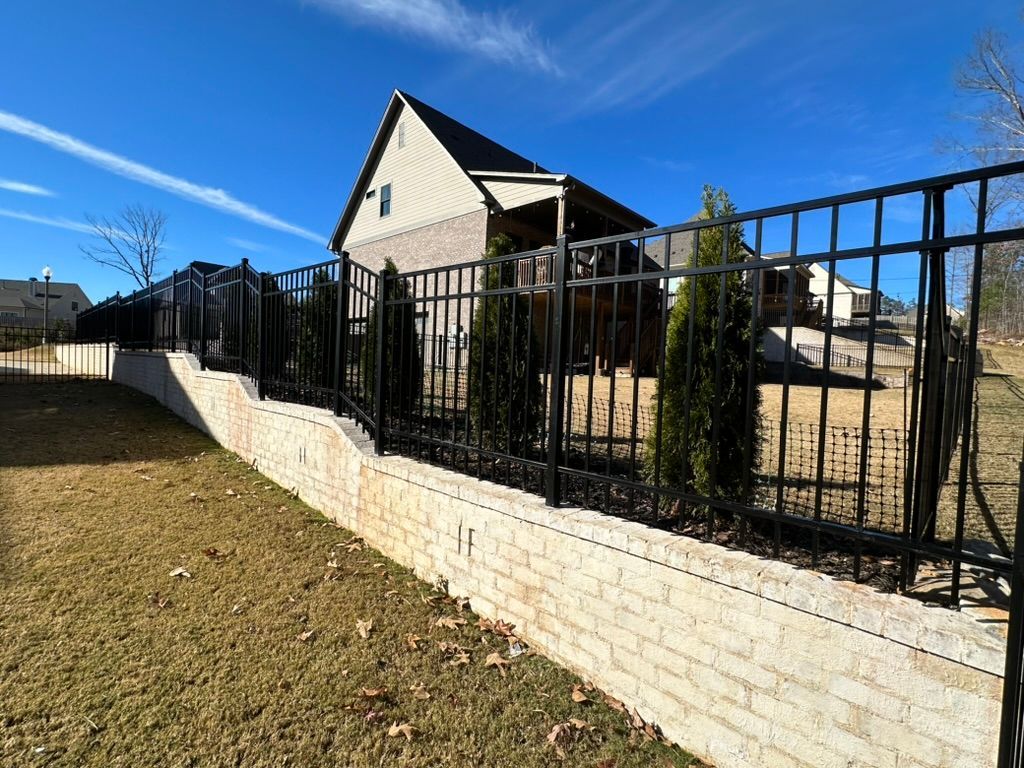 Black metal fence atop a beige retaining wall in front of a house on a sunny day.