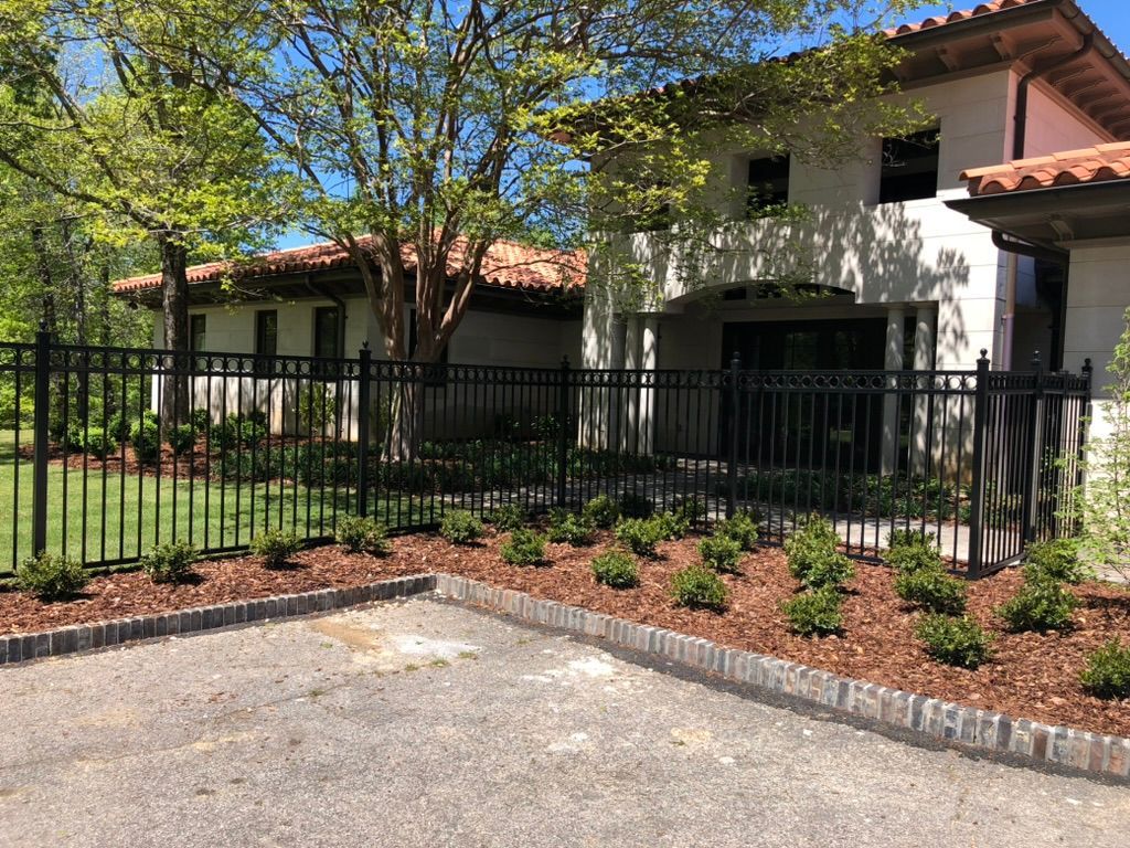 Black fence encloses a light-colored building with a terracotta roof, landscaped with small shrubs and mulch.