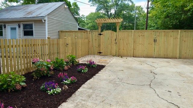 Wooden fence surrounds a yard with a garden in front. The gate is open.